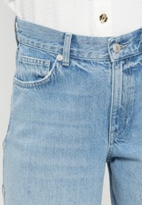 Jean en denim bleu clair à taille haute, avec une fermeture à boutons sur le devant, des poches latérales et des bords bruts le long des ouvertures de poche.