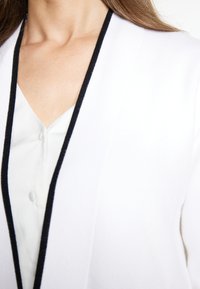 Cardigan bianco con bordi neri, dotato di scollatura a V. Tessuto liscio e camicia bianca con bottoni visibile sotto.