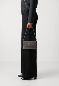 Sac à main gris métallique avec un design tressé, doté d'un rabat à pli et d'une anse en chaîne argentée. Porté avec un pantalon large noir et des talons.