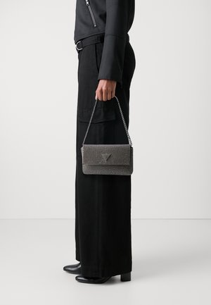 Sac à main gris métallique avec un design tressé, doté d'un rabat à pli et d'une anse en chaîne argentée. Porté avec un pantalon large noir et des talons.