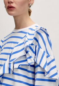 Witte blouse met blauwe horizontale strepen, voorzien van ruches op de schouders, een ronde halslijn en een zachte katoenen textuur.
