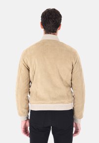 Giacca bomber in suede beige con colletto e polsini a coste, caratterizzata da una texture morbida e un design aderente. Sotto si indossano pantaloni neri.