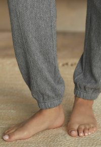 Joggers en tricot gris avec une texture côtelée et des poignets élastiques, présentés aux côtés d'un pied nu sur une surface tissée.