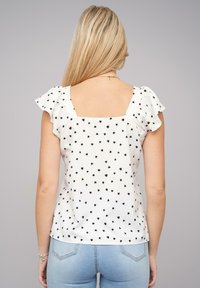Weiße Bluse mit kurzen Rüschärmeln, die ein schwarzes Herzmuster aufweist. Rechteckiger Rückendesign mit glatter Textur. Kombiniert mit hellblauen Jeans.