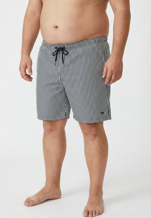 Mężczyzna w czarno-białych kąpielówkach w pionowe paski ze sznurkiem w talii, stoi boso na jednolitym tle.