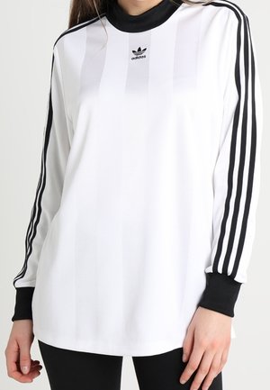 Valkoinen Adidas pitkähihaisen paita, jossa mustat kolmiraidat hihoissa sekä mustat kaulus ja kalvosimet, henkilö käyttää sitä.