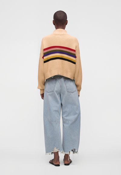 Personne vue de dos portant un pull beige à rayures colorées, un jean large bleu clair avec des ourlets effilochés et des sandales noires.