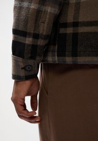 Brown checkered flannel shirt with black accents and a button cuff, worn over a tan pants. Textured fabric with visible stitching details.