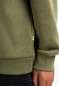 Groene sweatshirt van zacht materiaal, met geribbelde manchetten en onderzoom. Een klein groen label met een logo is zichtbaar op de zijnaad.