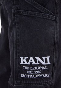 Tessuto denim nero con testo ricamato bianco "KANI THE ORIGINAL EST. 1989 REG.TRADEMARK" su una tasca, forma rettangolare con dettagli di cucitura.