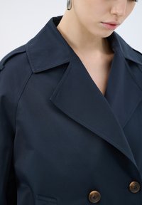 Marinblå dubbelskärmad trenchcoat med bred krage, texturerat tyg och bruna runda knappar. Nära vy av detaljer.