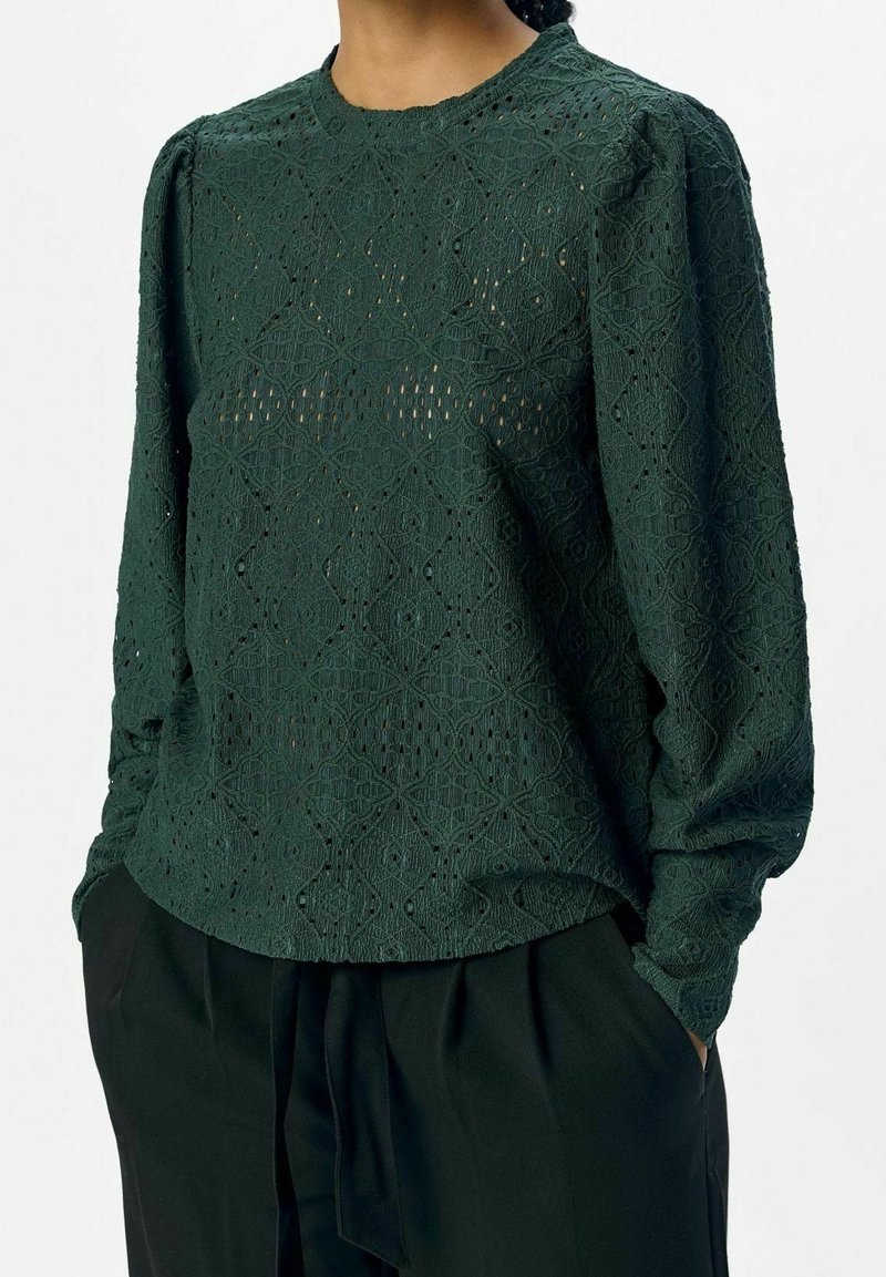 Femme portant un chemisier en dentelle vert foncé à manches longues avec un motif géométrique, assorti à un pantalon noir taille haute, les mains dans les poches.