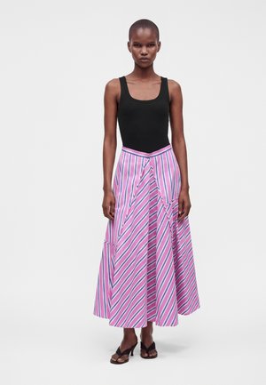 Vrouw draagt een zwarte mouwloze top, een lange roze-blauw gestreepte rok en zwarte open-teen sandalen met hak, staand tegen een effen achtergrond.