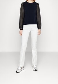 Haut navy avec des manches bouffantes transparentes à pois noirs, associé à un jean blanc évasé et des baskets grises avec des accents rouges.