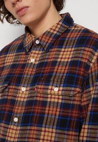 Chemise en flanelle dans un motif tartan marron, bleu et noir. Présente un col, des boutons de fermeture et deux poches poitrine. Fabriquée en tissu texturé.