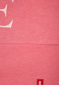 Koraalkleurige sweatshirt met geribbelde zoom. Kenmerkt zich door een textuurstof, grote witte print en een klein rood logo-label in de onderste hoek.