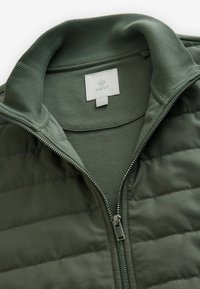 Grüne Zip-Jacke mit einem gerippten Kragen und gepolstertem Abschnitt. Verfügt über eine innere Schicht aus festem Stoff, mittlerem Gewicht und silbernem Reißverschluss.