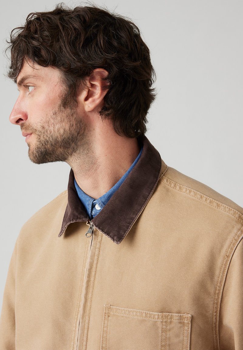 Chaqueta de lona color crema con un cuello de pana marrón. Cuenta con un cierre frontal y un bolsillo en el pecho. Lleva puesta una camisa de botones azul.