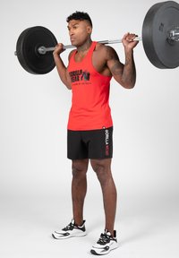 Rode tanktop met zwarte print, zwarte shorts met logo, en zwart-witte sneakers. Persoon die een halter heft met een geconcentreerde uitdrukking.