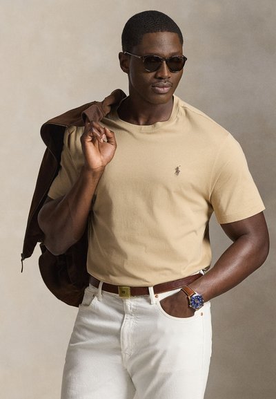 Mann mit Sonnenbrille und beigem T-Shirt trägt eine braune Jacke über der Schulter, mit weißen Hosen und einer Uhr mit blauem Zifferblatt am Handgelenk.
