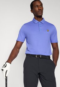 Vijolična polo majica z ovratnikom, z nežnim rumenim logotipom metulja. Kombinirana s črnimi hlačami in črnim tkanim pasom. Vidna bela golf rokavica.