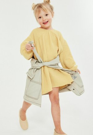 Jong meisje met blond haar in een gele jurk en een beige jasje om de taille geknoopt, glimlachend en een stap vooruit zet.