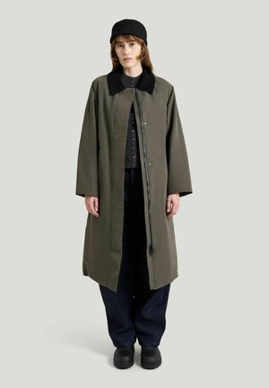 Ung person iført en lang olivengrøn frakke med sort krave, sort hue, sort knappet sweater, mørke bukser og sorte sko, der står oprejst.