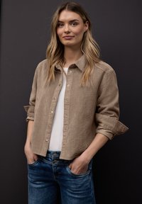 Jeune femme aux longs cheveux blonds portant une veste en velours côtelé beige sur une chemise blanche et un jean bleu, debout les mains dans les poches.