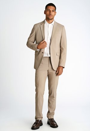 Beige Anzug, bestehend aus einer taillierten Jacke und passenden Hosen, kombiniert mit einem weißen Hemd mit Knöpfen, getragen mit dunkelbraunen Schuhen.