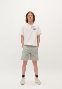 Witte polo met blauwe tekst "SCALPERS," lichtgrijze shorts en witte sneakers. Textuurstof, korte mouwen, casual pasvorm.