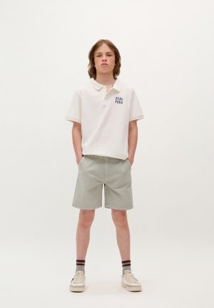 Polo bianco con testo blu "SCALPERS", pantaloni corti grigio chiaro e sneakers bianche. Tessuto texturizzato, maniche corte, vestibilità casual.