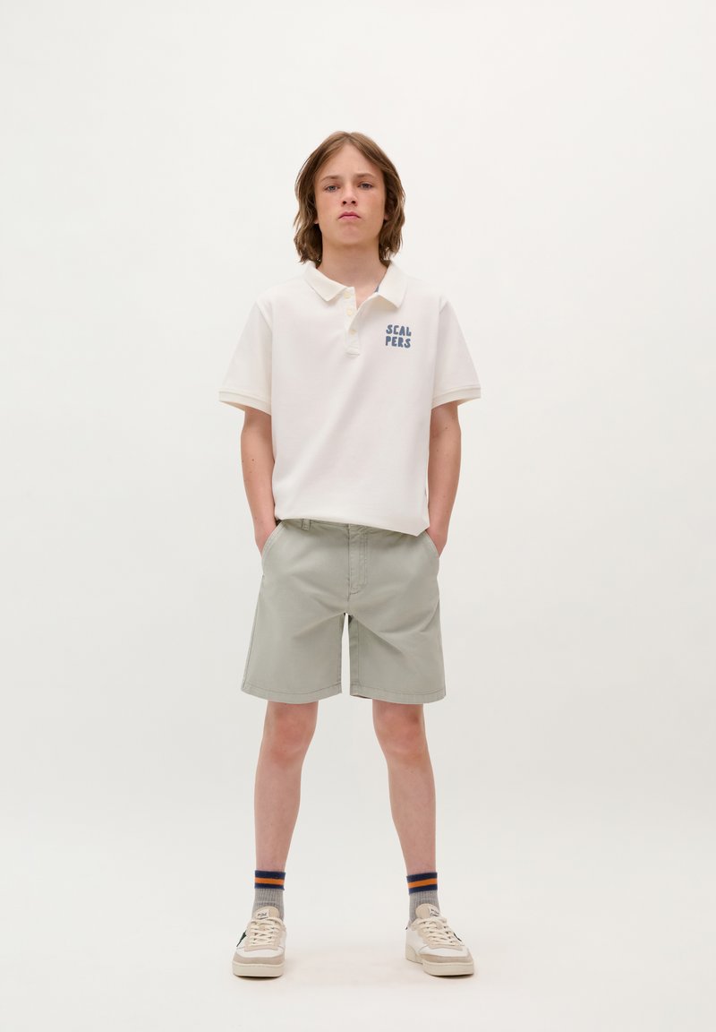 Witte polo met blauwe tekst "SCALPERS," lichtgrijze shorts en witte sneakers. Textuurstof, korte mouwen, casual pasvorm.