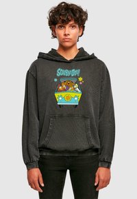 Čierna mikina s kapucňou vyrobená z mäkkej tkaniny, ktorá obsahuje farebnú grafiku "Scooby-Doo!" s postavami vo vnútri dodávky. Obsahuje prednú kapsu a kapucňu na sťahovanie.