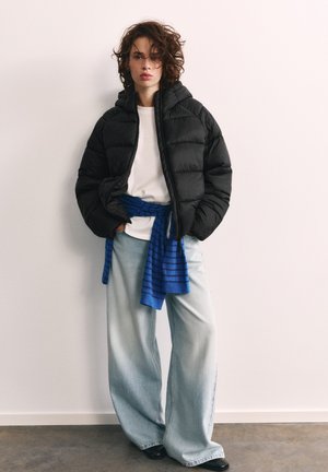 Personne aux cheveux bouclés portant une doudoune noire, une chemise blanche, un pull rayé bleu noué à la taille, un jean large bleu clair et des chaussures noires.