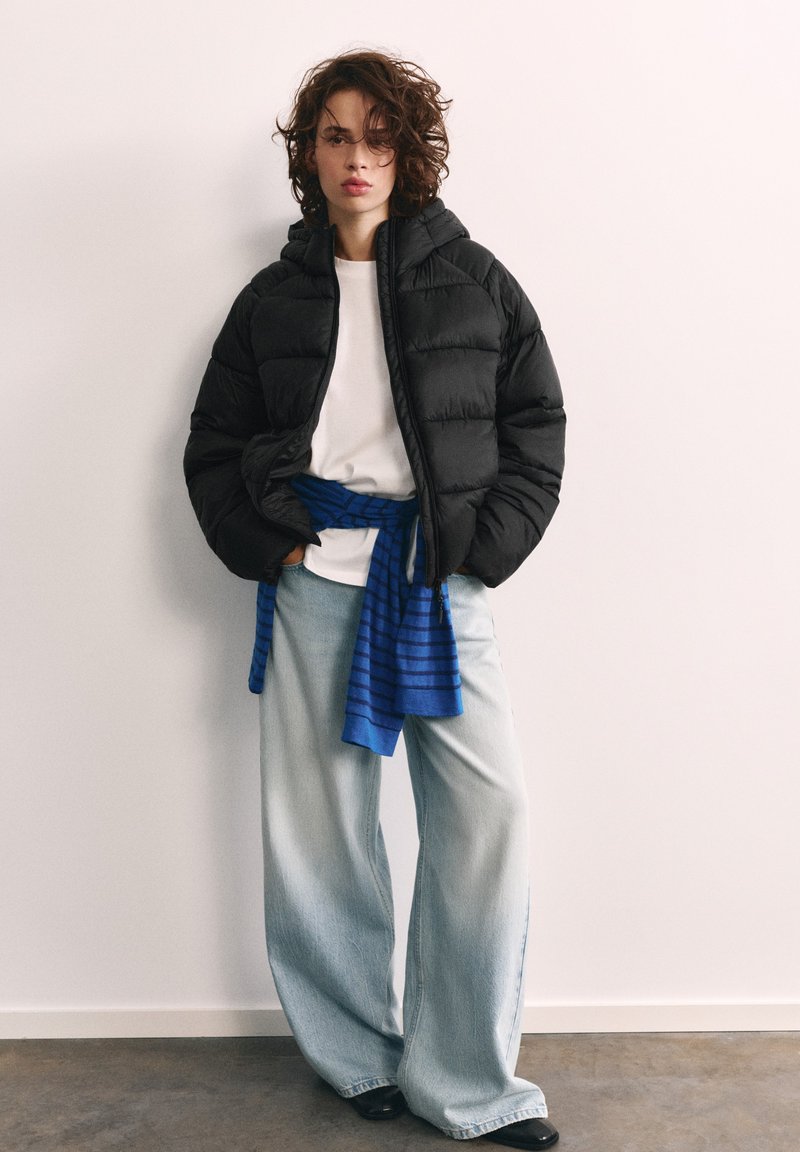 Personne aux cheveux bouclés portant une doudoune noire, une chemise blanche, un pull rayé bleu noué à la taille, un jean large bleu clair et des chaussures noires.