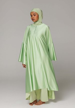 Vrouw die een lichtgroene satijnen abaya met wijde mouwen en een bijpassende hijab draagt, blootsvoets staand tegen een effen grijze achtergrond.