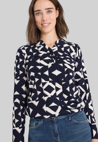 Camisa de manga larga azul marino con patrones geométricos blancos, con un cuello henley y tres botones, llevada sobre unos vaqueros azules.