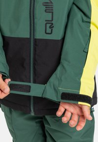 Ein Kind passt den Ärmel der grün, schwarz und gelb gestreiften Jacke mit einem Klettverschluss an und trägt passende grüne Hosen.