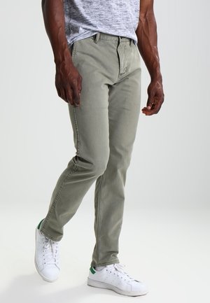 Uomo che indossa jeans grigio chiaro, sneakers bianche con dettagli verdi e una t-shirt grigia, in piedi con una gamba leggermente piegata.