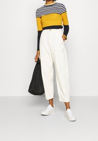 Gestreiftes Langarmoberteil in Gelb, Marineblau und Weiß; kombiniert mit hochgeschnittenen, cremefarbenen Hosen; weißen Sneakers und einer schwarzen Tasche.