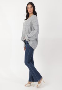 Grau oversized Pullover aus weichem Strick, mit V-Ausschnitt, überschnittenen Schultern und gerippten Bündchen, kombiniert mit dunklen Schlagjeans und weißen Absätzen.