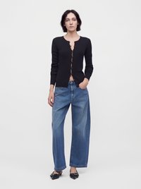 Femme portant un cardigan noir boutonné, un jean bleu à jambes larges et des chaussures pointues noires, debout sur un fond blanc uni.