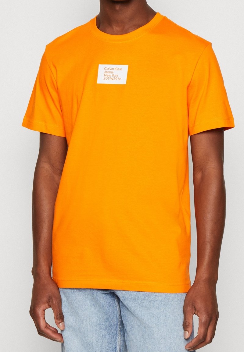 Hombre vistiendo una camiseta de manga corta de color naranja brillante con un logo cuadrado blanco que dice "Calvin Klein Jeans Nueva York 205 W 39 St." y jeans de color azul claro.
