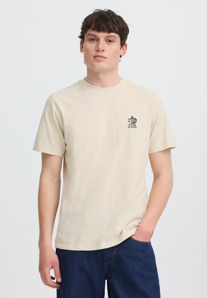 Ung mand iført en beige T-shirt med en lille sort kaffemaskine grafik på venstre bryst, kombineret med mørkeblå jeans, hånd i lommen.