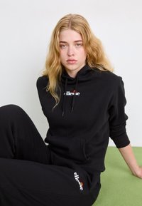 Sudadera con capucha negra y pantalones a juego de tejido suave. Cuenta con un bolsillo frontal y un logotipo en el pecho y el muslo.
