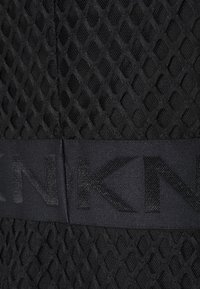 Tissu en maille noire avec un motif en forme de losanges, présentant une bande noire unie avec des lettres embossées. La surface texturée met en avant la tridimensionnalité.