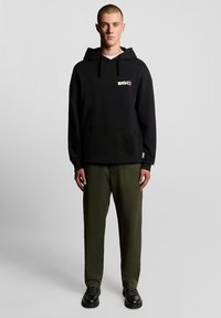 Juodas gobtuvu džemperis su priekine kišene, dekoruotas spalvingu logotipo elementu. Papildytas alyvuogių žalių kelnių ir juodų batų deriniu.