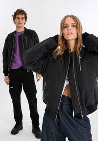 Black bomber jackets with ribbed cuffs, featuring zippered pockets and logos. The male model wears a purple shirt, and the female model has a white top.