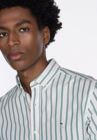 Camisa de algodón a rayas verdes y blancas con cuello abotonado y un pequeño logo bordado en el pecho. Textura suave, corte casual.