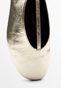 Metallisch goldener Slip-On-Schuh mit abgerundeter Spitze, glatter Textur und minimalen Nahtdetails. Das Innere ist schwarz und zeigt ein schlichtes Design.